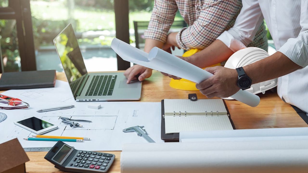 Deux professionnels en train d'examiner des plans de construction et de travailler sur un ordinateur portable, entourés d'outils techniques, d'une calculatrice, d'un cahier de notes, et d'un casque de sécurité sur une table de travail.